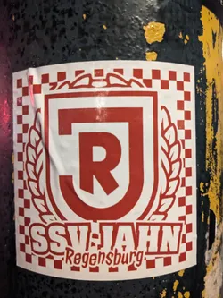 Street sticker A sticker featuring the logo of SSV Jahn Regensburg, a German football club. The logo is a red and white shield with the letters "J" and "R" in the center, surrounded by wheat ears. The text "SSV JAHN Regensburg" is written below the logo.