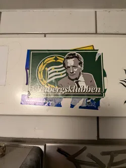 Street sticker The sticker features a stylized portrait of a man in a suit and tie, rendered in shades of gray and white. He is positioned against a dark green background. To the left of his head is a circular emblem with a yellow laurel wreath surrounding a flag with horizontal stripes in white and green, with the letters 'H' and 'F' visible. Below the portrait, the text 'Vrabergsklubben' is written in a stylized white font. At the bottom of the sticker, a blue strip contains the text 'WWW.MONTANA-CANS.COM'. The sticker appears to be slightly worn and has some torn edges.