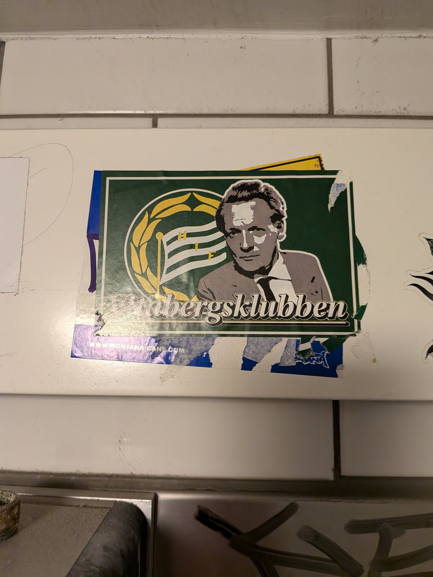 The sticker features a stylized portrait of a man in a suit and tie, rendered in shades of gray and white. He is positioned against a dark green background. To the left of his head is a circular emblem with a yellow laurel wreath surrounding a flag with horizontal stripes in white and green, with the letters 'H' and 'F' visible. Below the portrait, the text 'Vrabergsklubben' is written in a stylized white font. At the bottom of the sticker, a blue strip contains the text 'WWW.MONTANA-CANS.COM'. The sticker appears to be slightly worn and has some torn edges.