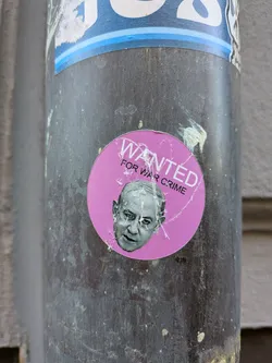 The sticker is circular with a vibrant pink background. In the upper portion, the word "WANTED" is prominently displayed in large, white, sans-serif capital letters with a slight curve. Below it, in smaller white capital letters, is the text "FOR WAR CRIME". Centered below the text is a grayscale portrait of an elderly man with a stern, displeased expression. The sticker shows signs of wear and tear, with visible creases and peeling edges, indicating it has been affixed to the surface for some time. Some white and light blue elements from another sticker are visible above it.