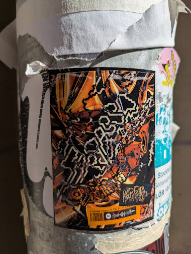 A close-up shot shows an abstract sticker with a geometric pattern of orange and black. The bottom of the sticker shows a Spotify code and the text reads "KELP FOREST". Another text on the sticker says "RIPFACE". The sticker is stuck on a pole and appears to be part of a collection of other stickers.