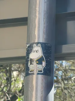 Street sticker The sticker features a white, hippopotamus-like cartoon character, strongly resembling Moomintroll, with a grumpy or determined expression. It has large, round eyes with black pupils and thick eyebrows furrowed downwards. The character is wearing a dark blue (or black) sports jersey with a yellow collar and yellow stripes on the sleeves and chest. Below the jersey, it wears light grey shorts or pants and dark blue shoes with yellow trim. The background of the sticker is a dark blue with a speckled, distressed white pattern, giving it a somewhat textured or starry appearance. The sticker itself shows signs of wear and tear, particularly in the center where it is peeling and torn, obscuring part of the character's midsection.