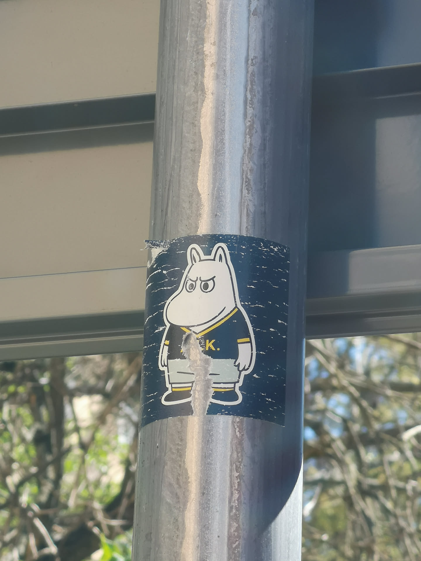 The sticker features a white, hippopotamus-like cartoon character, strongly resembling Moomintroll, with a grumpy or determined expression. It has large, round eyes with black pupils and thick eyebrows furrowed downwards. The character is wearing a dark blue (or black) sports jersey with a yellow collar and yellow stripes on the sleeves and chest. Below the jersey, it wears light grey shorts or pants and dark blue shoes with yellow trim. The background of the sticker is a dark blue with a speckled, distressed white pattern, giving it a somewhat textured or starry appearance. The sticker itself shows signs of wear and tear, particularly in the center where it is peeling and torn, obscuring part of the character's midsection.