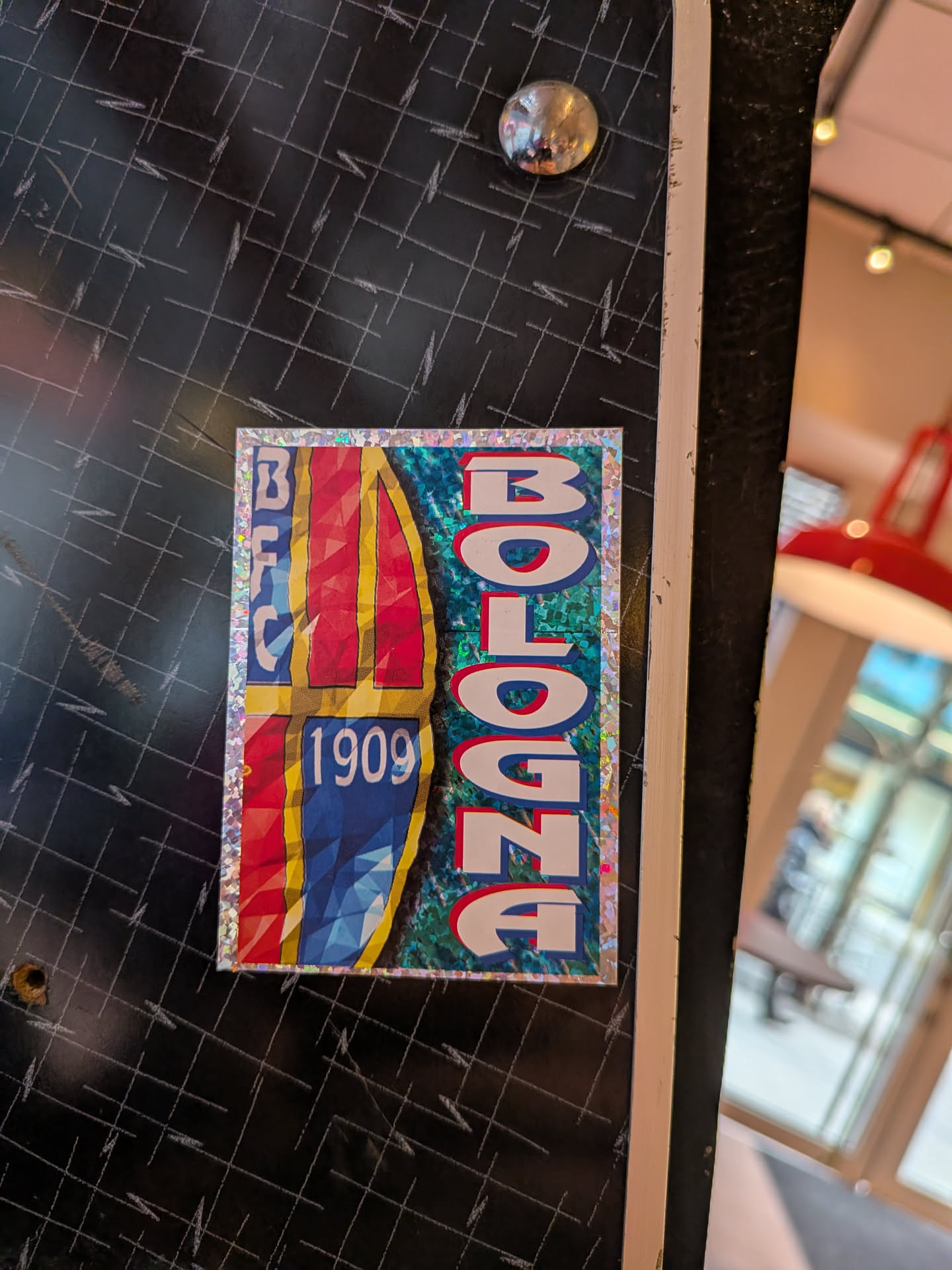 A vertically oriented rectangular sticker featuring a shimmering holographic border. The left side of the sticker displays a stylized crest or shield design with vertical stripes in red, yellow, and blue, rendered with a faceted or low-polygon effect. Stacked vertically on the far left of this section are the letters 'BFC' in white with a blue outline, and below them, '1909' in the same style, positioned over the blue stripe. The right side of the sticker features the word 'BOLOGNA' written vertically from top to bottom in large, bold white letters with a red outline. This text is set against a textured, dynamic background of blues and greens, which appears to have a pixelated or water-like pattern.