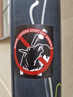Street sticker A round sticker with a red circle and a line through it. Inside the circle is a drawing of a cow being slaughtered. The text around the circle says "Animal lovers don't eat animals".