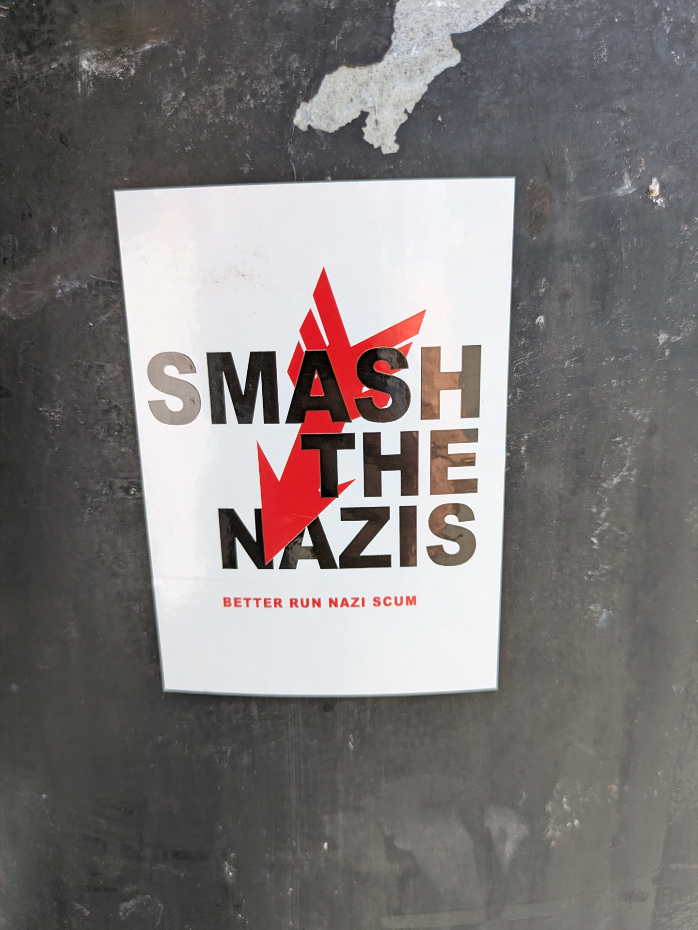 The sticker is white with a red starburst design cutting through the words "SMASH THE NAZIS" written in a bold, sans-serif font. Beneath this, in smaller red text, it reads "BETTER RUN NAZI SCUM". The sticker is stuck on a dark, possibly metal, surface.