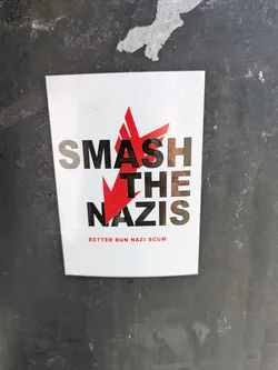 Street sticker The sticker is white with a red starburst design cutting through the words "SMASH THE NAZIS" written in a bold, sans-serif font. Beneath this, in smaller red text, it reads "BETTER RUN NAZI SCUM". The sticker is stuck on a dark, possibly metal, surface.