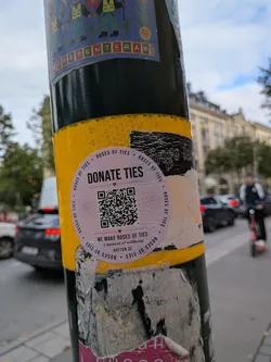 Street sticker A circular, light-pink sticker affixed to a yellow and black pole. The sticker features the bold text 'DONATE TIES' at the top, a central QR code flanked by two faint rose line drawings, and the text 'WE MAKE ROSES OF TIES', 'A moment of wellbeing', and 'ARTTEN.SE' at the bottom. The outer border is lined with the repeating phrase 'ROSES OF TIES'.