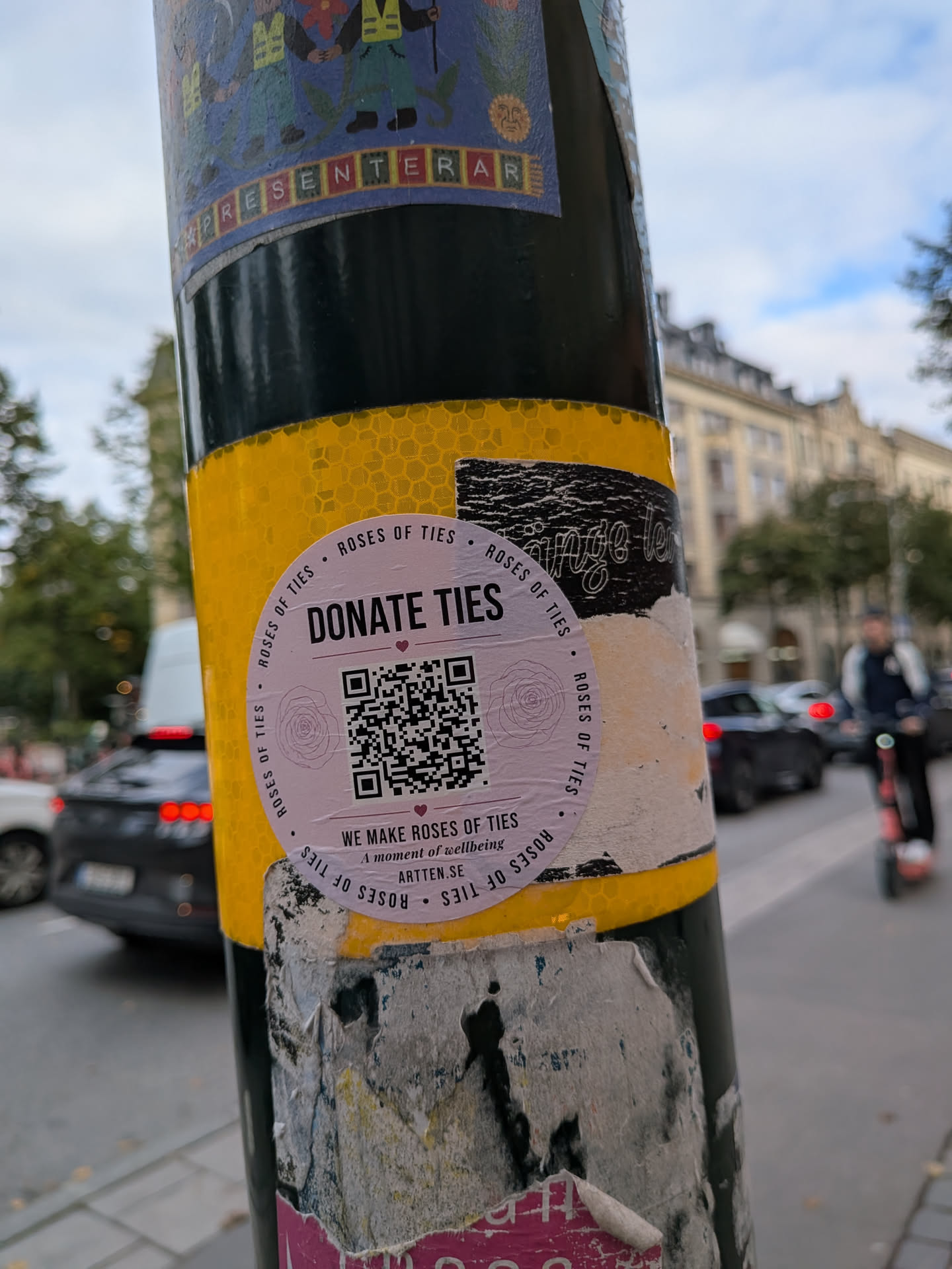 A circular, light-pink sticker affixed to a yellow and black pole. The sticker features the bold text 'DONATE TIES' at the top, a central QR code flanked by two faint rose line drawings, and the text 'WE MAKE ROSES OF TIES', 'A moment of wellbeing', and 'ARTTEN.SE' at the bottom. The outer border is lined with the repeating phrase 'ROSES OF TIES'.