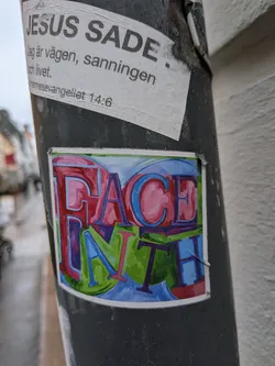 Street sticker The sticker is rectangular with a white border, featuring the words "FACE" and "FAITH" rendered in a vibrant, graffiti-inspired bubble letter style. The word "FACE" is positioned above "FAITH". "FACE" is primarily in shades of red and pink with blue outlines and shadows, creating a three-dimensional effect. "FAITH" is predominantly in shades of green with blue and purple outlines and shadows, also exhibiting a strong 3D appearance. The letters are large and bold, filling the sticker's frame, and are slightly intertwined, giving the composition a dynamic and energetic feel. The background behind the letters appears to be a blend of light blue and purple.