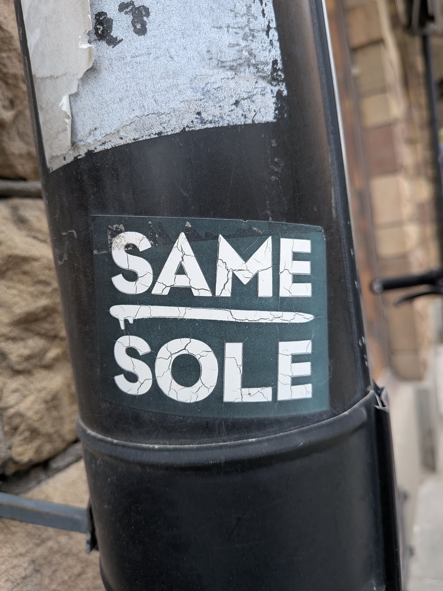 The sticker is rectangular with a dark teal or dark green background. It features two lines of bold, sans-serif text in white. The top line reads 'SAME' and the bottom line reads 'SOLE'. Between the two words, there is a horizontal line with a small drip-like element extending downwards from its center. The white text and the line are heavily cracked, giving them a distressed or aged appearance, suggesting wear and time. The edges of the sticker are slightly worn and dirty, adhering to a dark, possibly metal, pole.
