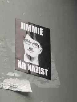 Street sticker A rectangular, vertically oriented sticker with a dark background featuring a stylized, grayscale portrait of a person wearing glasses and a toothbrush mustache, strongly resembling Adolf Hitler. Above the portrait, in bold white capital letters, is the word 'JIMMIE'. Below the portrait, also in bold white capital letters, are the words 'ÄR NAZIST'. The sticker appears slightly worn and adhered to a gray wall.