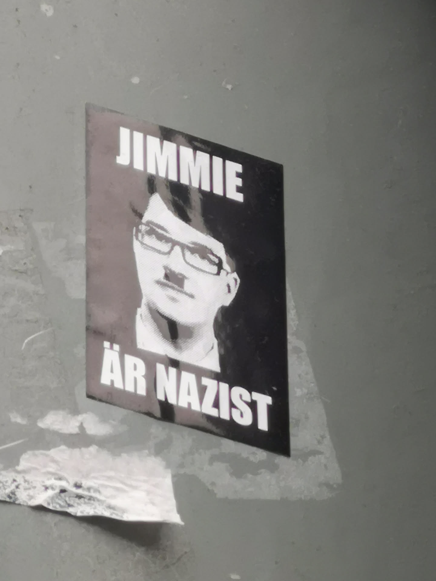 A rectangular, vertically oriented sticker with a dark background featuring a stylized, grayscale portrait of a person wearing glasses and a toothbrush mustache, strongly resembling Adolf Hitler. Above the portrait, in bold white capital letters, is the word 'JIMMIE'. Below the portrait, also in bold white capital letters, are the words 'ÄR NAZIST'. The sticker appears slightly worn and adhered to a gray wall.
