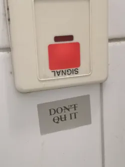 Street sticker A rectangular sticker, light gray in color, featuring the motivational phrase 'DON'T QUIT' in dark gray or black serif font. 'DON'T' is on the top line, and 'QUIT' is on the bottom line, centered on the sticker. The sticker is applied to a white tiled surface, positioned below a beige electrical or alarm panel.