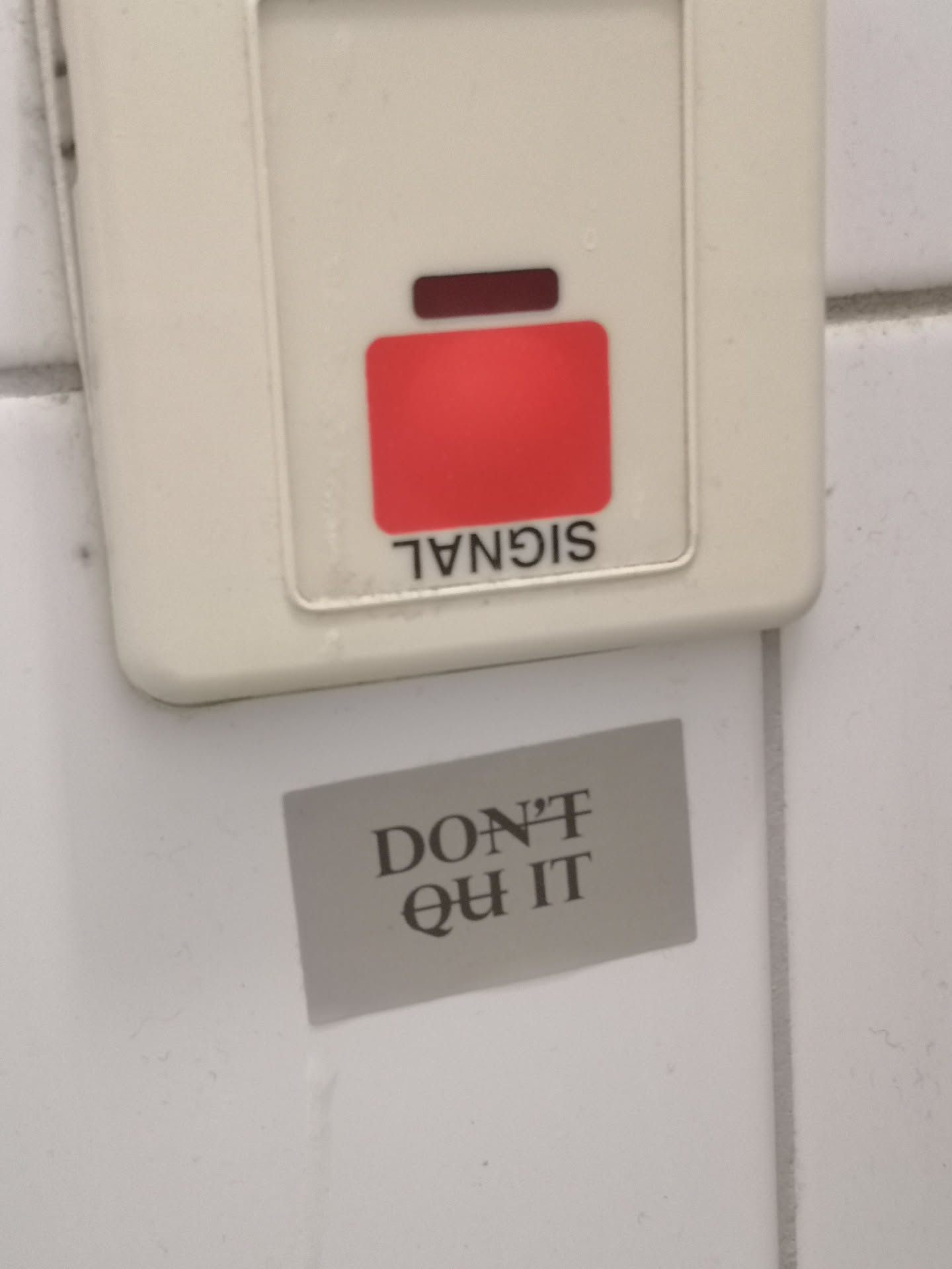 A rectangular sticker, light gray in color, featuring the motivational phrase 'DON'T QUIT' in dark gray or black serif font. 'DON'T' is on the top line, and 'QUIT' is on the bottom line, centered on the sticker. The sticker is applied to a white tiled surface, positioned below a beige electrical or alarm panel.