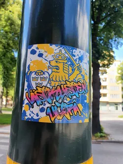 Street sticker A colorful sticker featuring the Swedish coat of arms lion in a graffiti style. The background is a mix of blue and yellow splotches. The text below the lion appears to say "VÄSTERMÄSTEN LILLA VITA", though it is somewhat damaged.