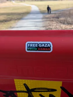 The sticker is a rectangular design with rounded corners and a prominent white border. The main background of the sticker is black. The text 'FREE GAZA' is displayed in bold white capital letters on the top line. Below it, the text 'FROM HAMAS' is presented, with 'FROM' in white capital letters and 'HAMAS' in green capital letters, featuring a subtle red outline or shadow, reminiscent of the Palestinian flag colors. The sticker is affixed to a textured, bright red surface.