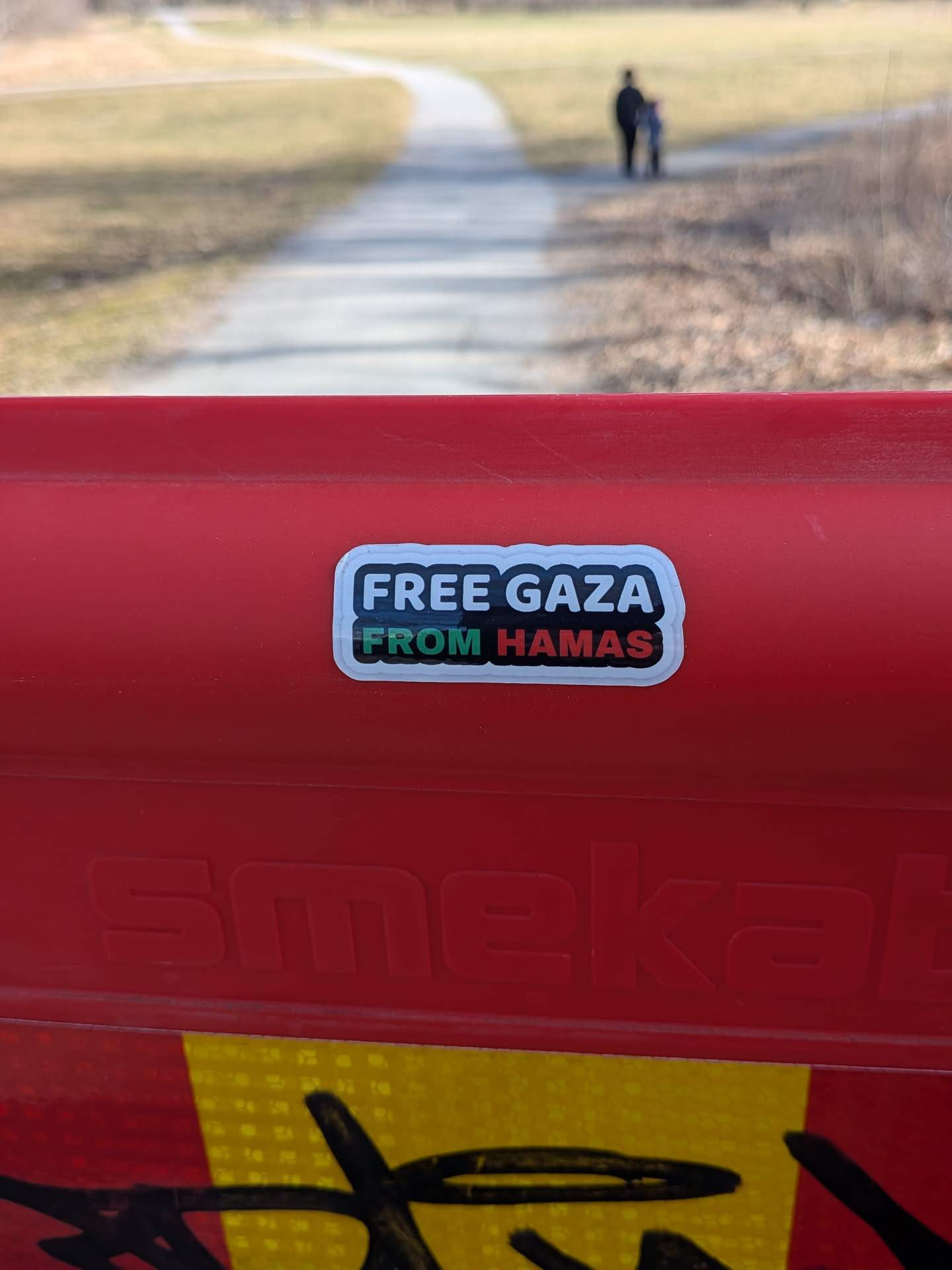 The sticker is a rectangular design with rounded corners and a prominent white border. The main background of the sticker is black. The text 'FREE GAZA' is displayed in bold white capital letters on the top line. Below it, the text 'FROM HAMAS' is presented, with 'FROM' in white capital letters and 'HAMAS' in green capital letters, featuring a subtle red outline or shadow, reminiscent of the Palestinian flag colors. The sticker is affixed to a textured, bright red surface.
