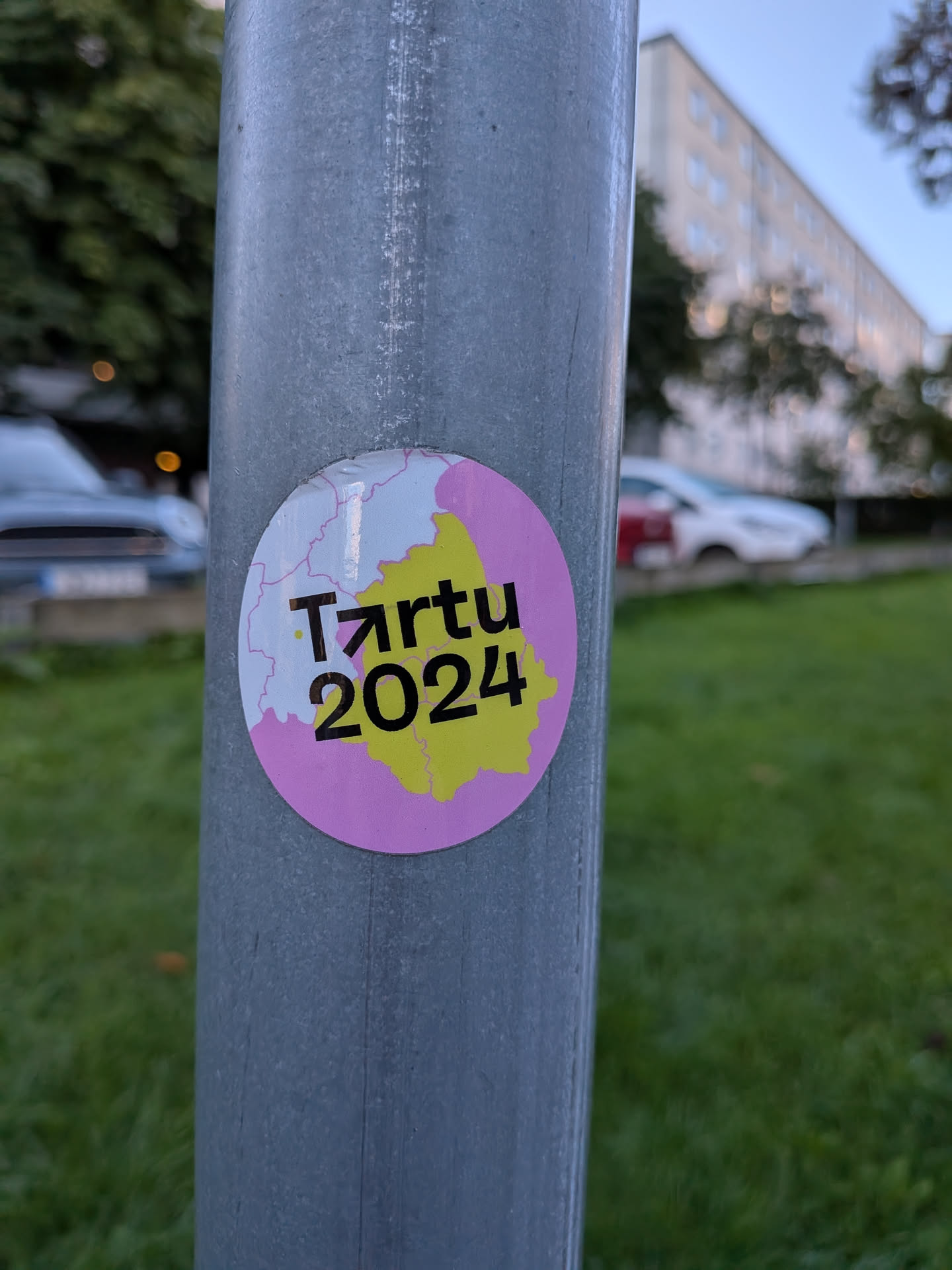 The sticker is circular with a light pink border. The center features a stylized map outline in white, with pink accents. Inside the map outline, the text 'Tartu' is written in a dark, bold font, with an upward-pointing arrow replacing the 'A'. Below 'Tartu', the number '2024' is printed in a similar dark, bold font. The map area within the border is filled with a bright yellow color. The overall design is clean and modern, suggesting an event or campaign.