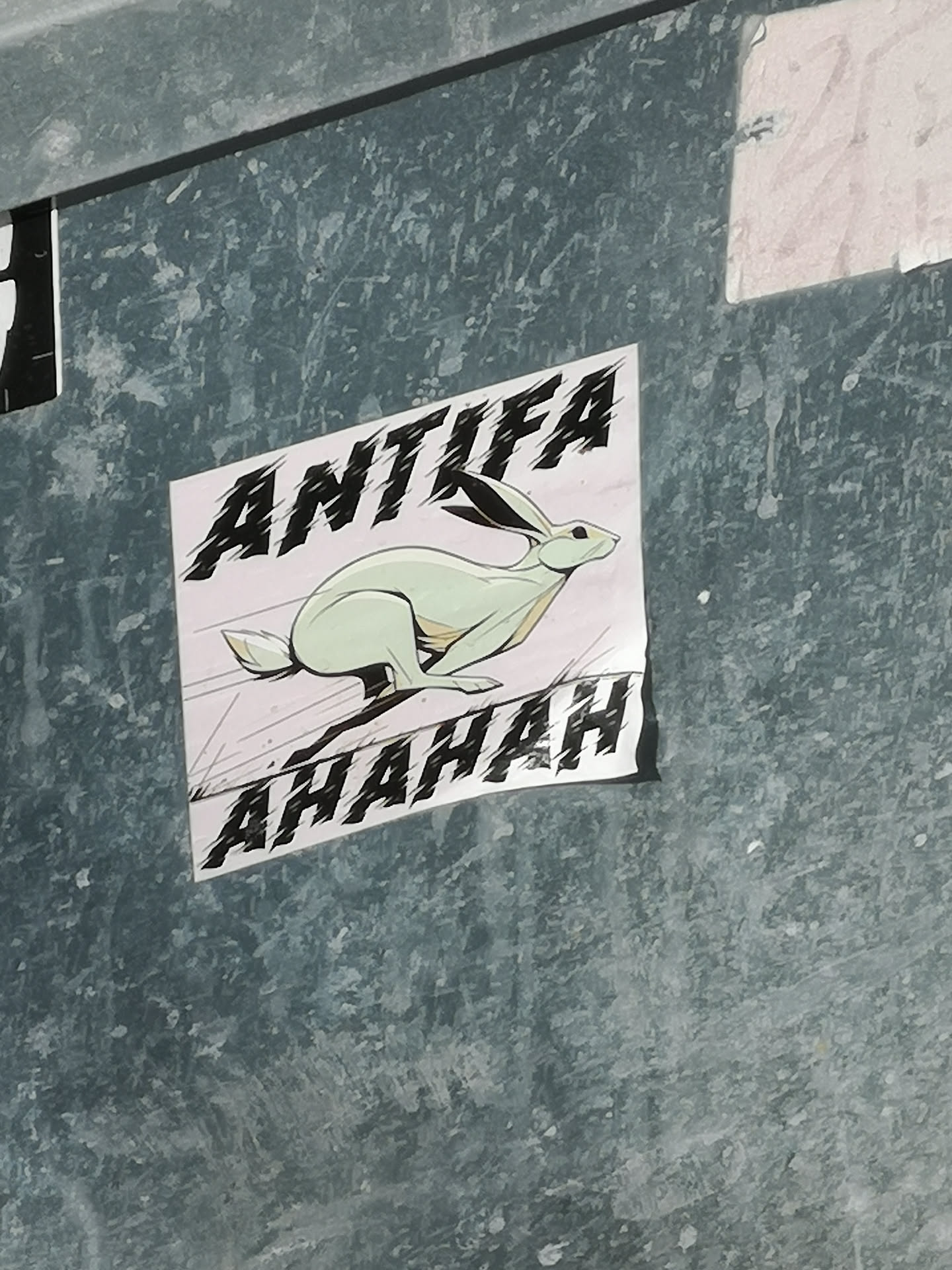 The sticker features a stylized, light green or mint-colored rabbit (or hare) depicted in a dynamic running pose, facing right. It has black outlines and subtle yellow-brown accents. Above the rabbit, the word "ANTIFA" is written in large, bold, black letters with a distressed, jagged-edge font. Below the rabbit, the word "AHAHAH" is written in a similar style. The background of the sticker is a plain off-white or very light grey, contrasting with the dark, textured surface it's adhered to. The sticker is rectangular and placed diagonally.