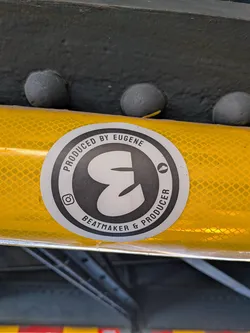 Street sticker The sticker is circular with a white border. The inner design features a stylized, bold 'E' in black, resembling a sound wave or a musical note. Around the 'E', in a circular band, is text in black capital letters. The top arc reads 'PRODUCED BY EUGENE', and the bottom arc reads '& PRODUCER'. To the left of the 'E' is a small Instagram logo, and to the right is a small cloud icon. The background of the inner circle is black.