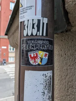 Street sticker The sticker features a black background with a repeating pattern of eagles. At the top, in white block letters outlined in black, is the text 'DIE MACHT DER'. Below this, in larger white block letters with a black outline, is 'SEENPLATTE'. The central element is a shield divided into three parts. The left section depicts a red eagle on a white background. The middle section shows a blue and white wavy pattern, a golden crown, and a black half-face with a golden helmet. The right section again shows a red eagle on a white background. Below the shield, the text 'SV BURG STARGARD 09' is visible in white letters. The shield itself is framed by a yellow border.