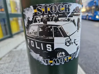 A sticker featuring a vintage police car, with text suggesting opposition to the police.  The sticker is affixed to a dark green pole, and appears worn and partially peeled.