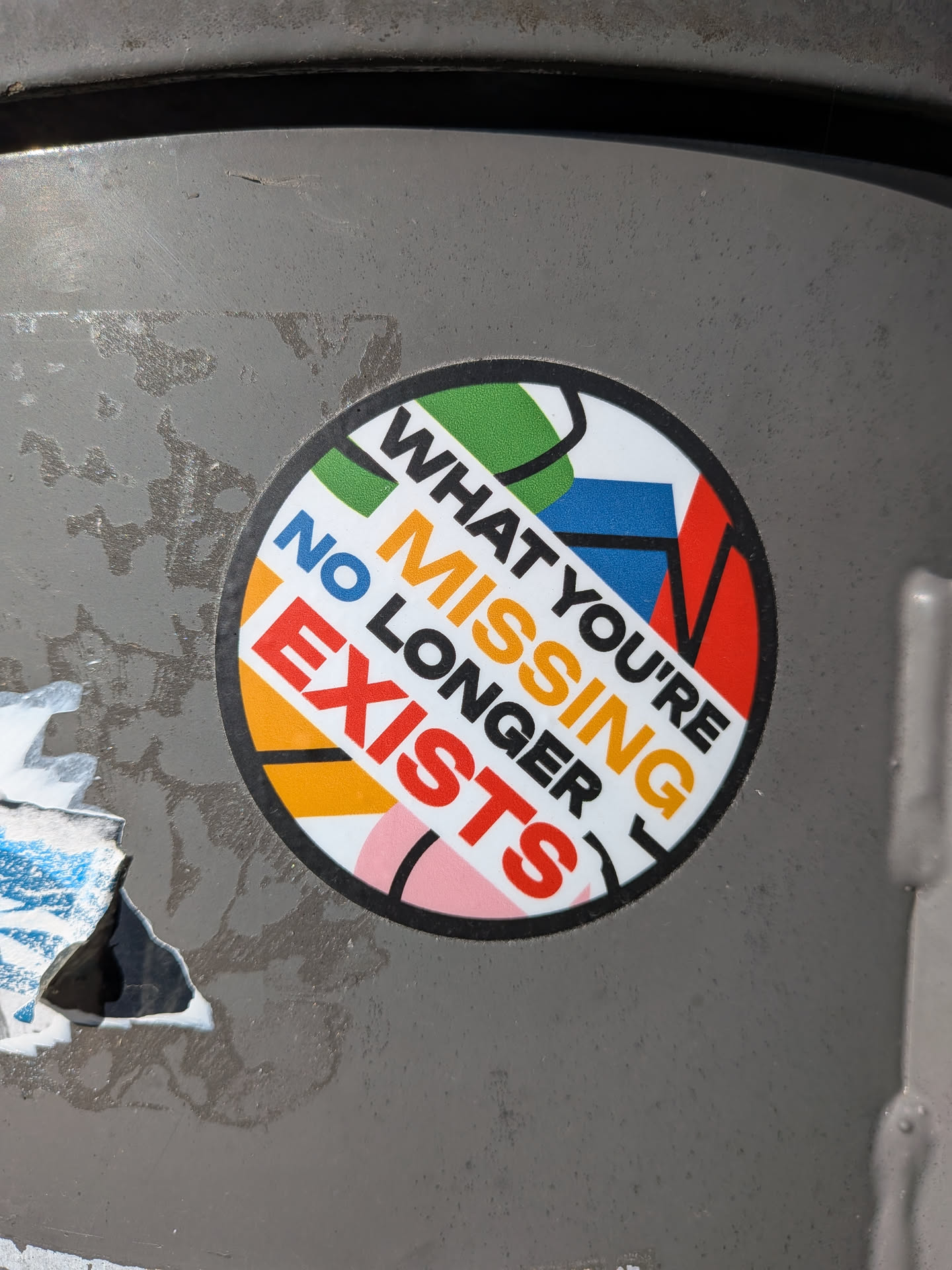 The circular sticker features the text "WHAT YOU'RE MISSING NO LONGER EXISTS" printed in bold, sans-serif fonts. The text is arranged in a diagonal, slightly curved manner across the sticker. The background is a collage of abstract geometric shapes in vibrant colors, including green, blue, red, orange, yellow, and pink, against a white base. A thick black border encircles the entire design.