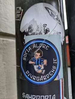 A round sticker featuring a cartoon illustration of a soccer player with the text "Sofia Äldre Djurgårdens IF" around the illustration. The sticker has a black background with light blue accents.