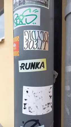Street sticker A small, rectangular sticker with the word "RUNKA" written in a stylized, rainbow-like font. The sticker has a slightly worn look, showing some signs of being affixed to a surface for some time. It's one among several stickers and graffiti on a gray pole.