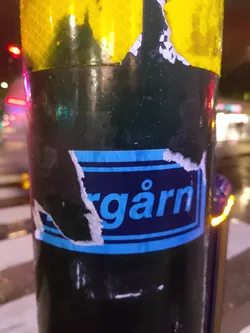 A partially damaged blue and dark blue sticker with the word "bergärn" visible.  It's affixed to a dark-colored pole outdoors at night. The sticker shows some wear and tear with pieces missing around the edges. The background is blurry, showing a nighttime urban scene.