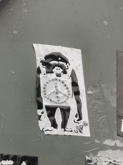 Street sticker The sticker features a black and white illustration of an upright chimpanzee or ape. The ape wears a dark beret adorned with a small white star, reminiscent of revolutionary figures. Over its chest, it wears a light-colored vest or bib, on which a large peace symbol is depicted, formed entirely by a chain of small, stylized flowers. Inside the floral peace symbol, text is visible. At the bottom right, near the ape's foot, a small drawing of a single flower with leaves is present. The overall style is high-contrast, resembling a stencil or woodcut. The sticker itself is a rectangular piece of white paper, showing slight wear and curled edges, adhered to a textured grey wall.
