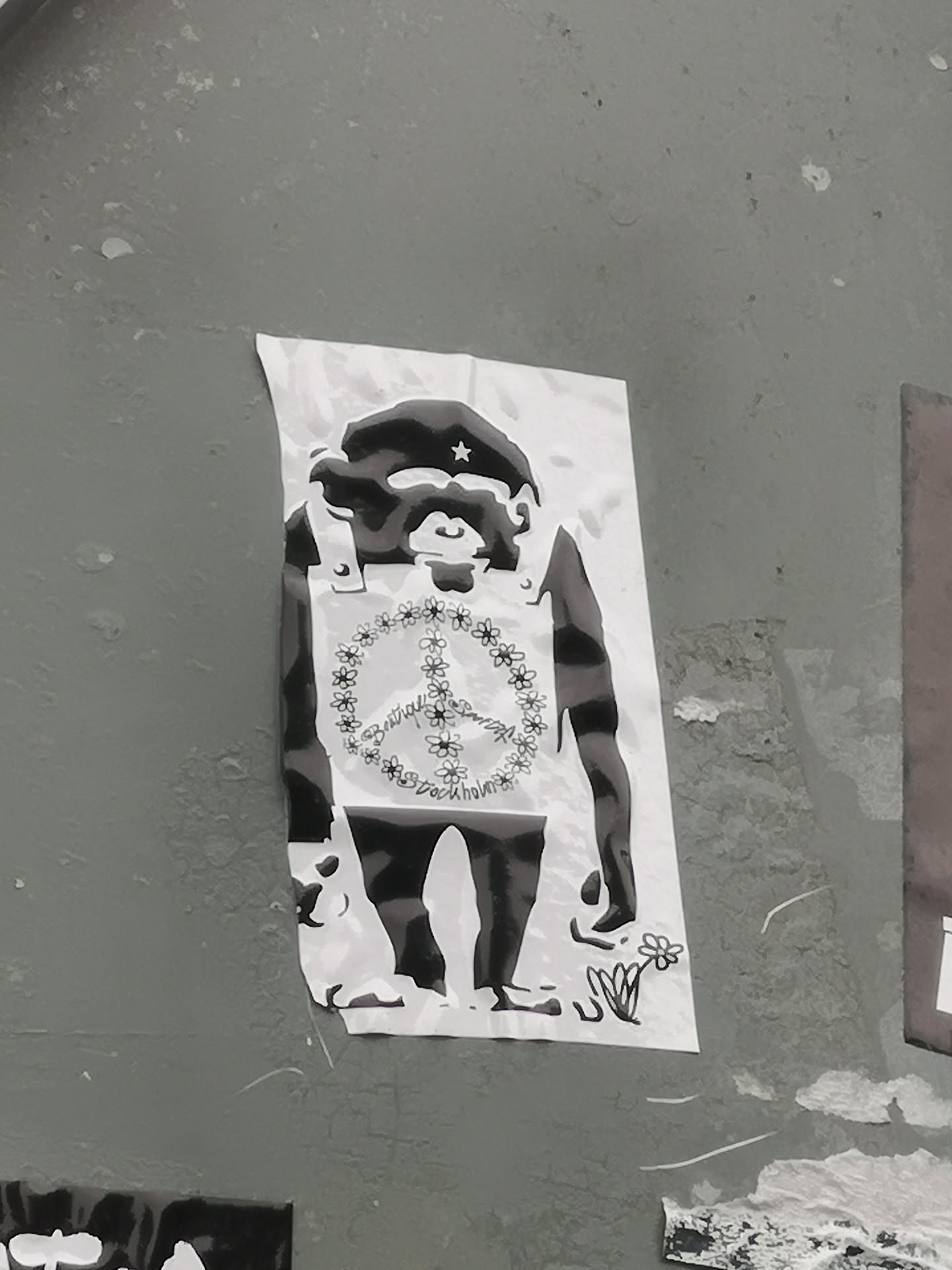 The sticker features a black and white illustration of an upright chimpanzee or ape. The ape wears a dark beret adorned with a small white star, reminiscent of revolutionary figures. Over its chest, it wears a light-colored vest or bib, on which a large peace symbol is depicted, formed entirely by a chain of small, stylized flowers. Inside the floral peace symbol, text is visible. At the bottom right, near the ape's foot, a small drawing of a single flower with leaves is present. The overall style is high-contrast, resembling a stencil or woodcut. The sticker itself is a rectangular piece of white paper, showing slight wear and curled edges, adhered to a textured grey wall.