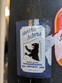 Street sticker The sticker is predominantly white with a decorative blue border featuring ornate scrollwork and a chain-like pattern. In the center, a black bear with red-clawed paws is depicted in a dynamic pose, with its mouth open showing teeth. The bear is outlined by thin grey radiating lines. Above the bear, in stylized blue cursive lettering, is 'Hertha Inters'. Below the bear, in block lettering, it reads 'Berlin's finest INTERNATIONAL IMPORTS.', with a small silhouette of a park bench beneath the word 'INTERNATIONAL'. At the bottom, centered, is '• SEIT 2022 •' in a smaller font.