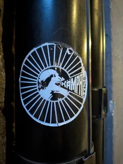 Street sticker The sticker is circular and predominantly black and white. It features a graphic illustration of a face in profile, with its mouth open as if shouting or speaking. The face is surrounded by a radial pattern of thin white lines emanating from the center, resembling sun rays or a burst. The text 'KAMPF' is prominently displayed in bold, blocky white letters within the right half of the circular design, integrated with the radial lines. The sticker appears to be slightly worn and peeling at the edges, indicating it has been applied to a surface for some time.