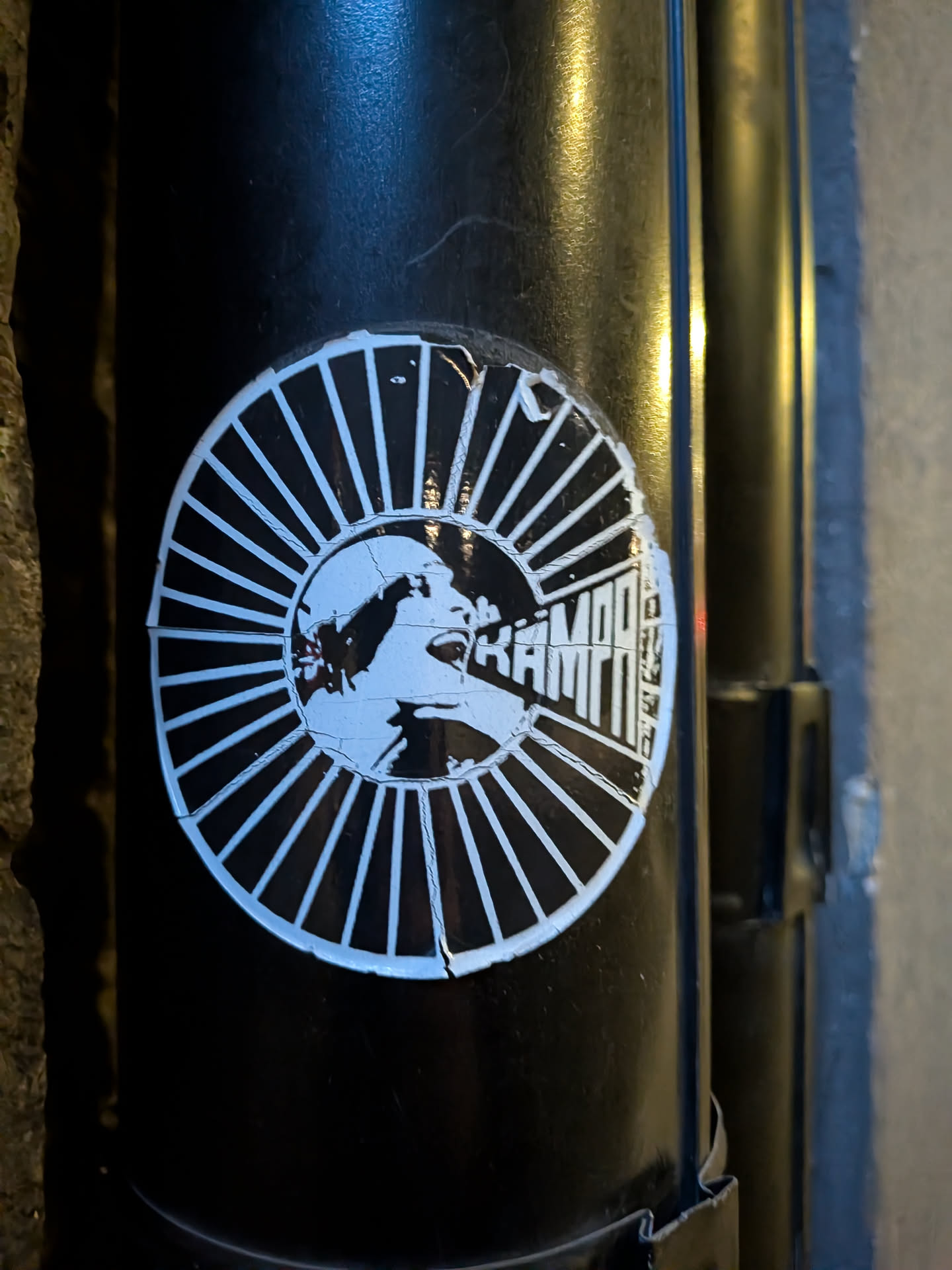 The sticker is circular and predominantly black and white. It features a graphic illustration of a face in profile, with its mouth open as if shouting or speaking. The face is surrounded by a radial pattern of thin white lines emanating from the center, resembling sun rays or a burst. The text 'KAMPF' is prominently displayed in bold, blocky white letters within the right half of the circular design, integrated with the radial lines. The sticker appears to be slightly worn and peeling at the edges, indicating it has been applied to a surface for some time.