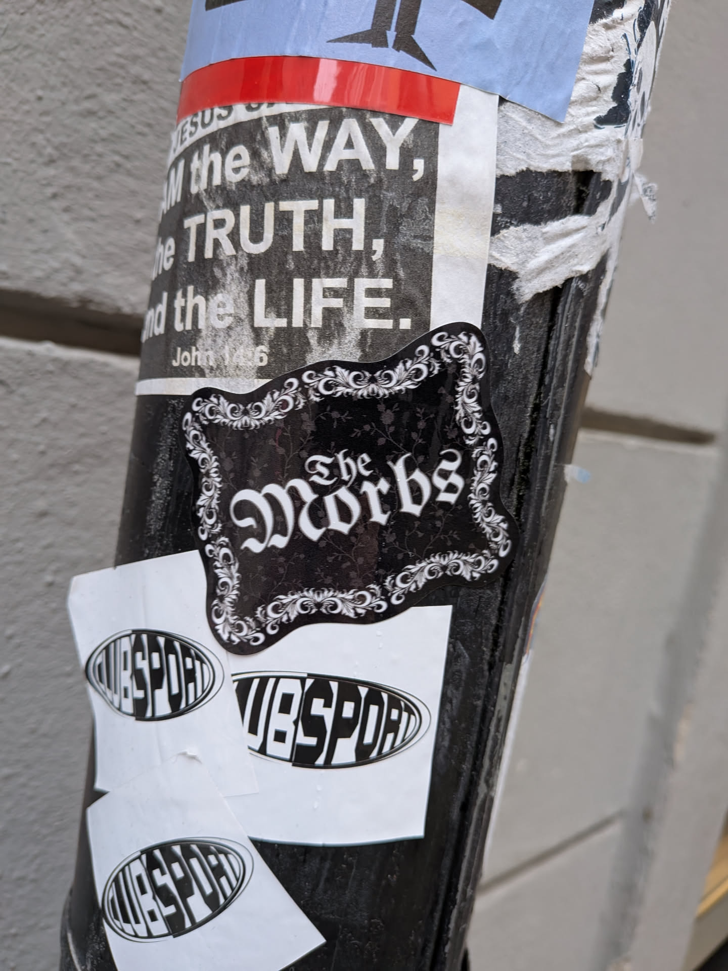 The sticker is an irregularly shaped, ornate design featuring a black background with white text and a decorative white border. The text 'The Morbs' is prominently displayed in a stylized, gothic-inspired font, with 'The' appearing smaller and positioned above 'Morbs'. The black background subtly incorporates a dark, almost invisible, floral or damask pattern. The white border is composed of intricate scrollwork and floral motifs, giving the sticker a vintage or elegant gothic aesthetic.