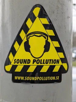 Street sticker A triangular yellow and black sticker with a person wearing earmuffs in the center and the text 'SOUND POLLUTION' and 'WWW.SOUNDPOLLUTION.SE'.