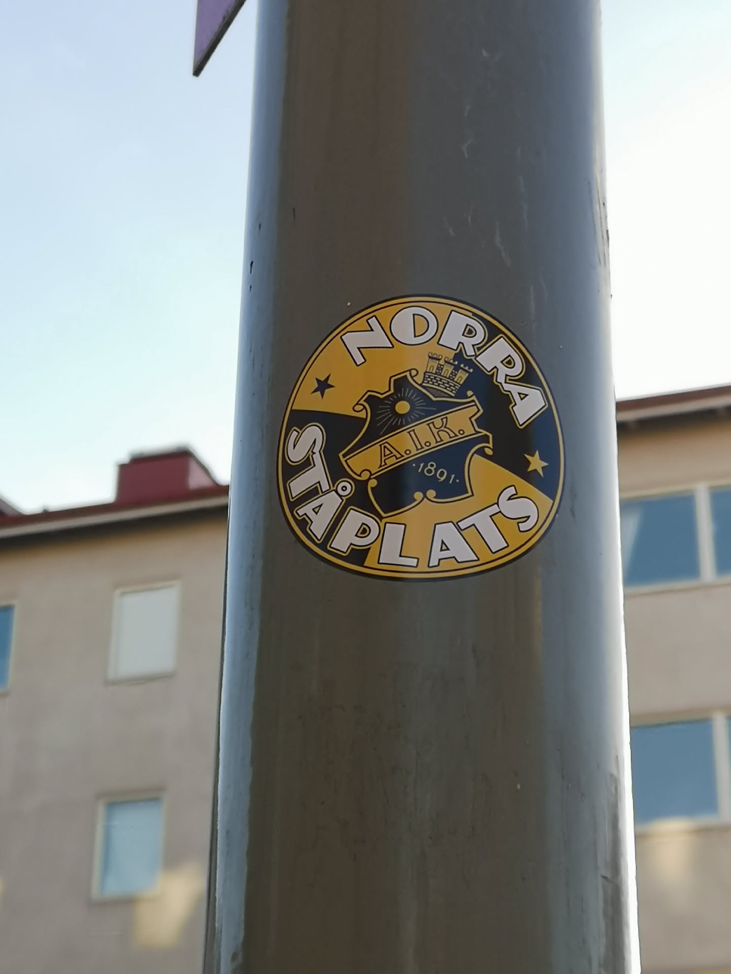 A circular sticker predominantly in yellow and black, featuring a central crest. The crest is black with yellow details, including a stylized sun, a crown, and the letters 'A.I.K.' with the year '1891' beneath them. Surrounding the crest, along the top curve, is the word 'NORRA' in white capital letters with a black outline, set against a yellow background. Along the bottom curve, also in white capital letters with a black outline on a yellow background, is 'STÅPLATS'. Two yellow stars are positioned on the yellow background, one on each side, separating the 'NORRA' and 'STÅPLATS' text.