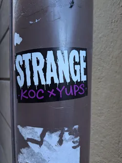 Street sticker A rectangular sticker with a solid black background. The word "STRANGE" is prominently displayed in large, bold, white capital letters, featuring a dripping paint effect at the bottom of each character. Below this, in smaller, purple, handwritten-style capital letters, is the text "KOC x YUPS.", flanked by small white dots. The sticker is applied to a brown pole, showing some wear and tear, with remnants of other stickers visible below it.