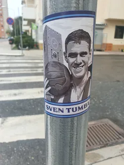Street sticker A grayscale sticker featuring a portrait of Sven Tumba, a Swedish ice hockey player, holding a soccer ball.  A building is depicted in the background. The sticker has a light blue border with the name "SVEN TUMBA" printed at the bottom.