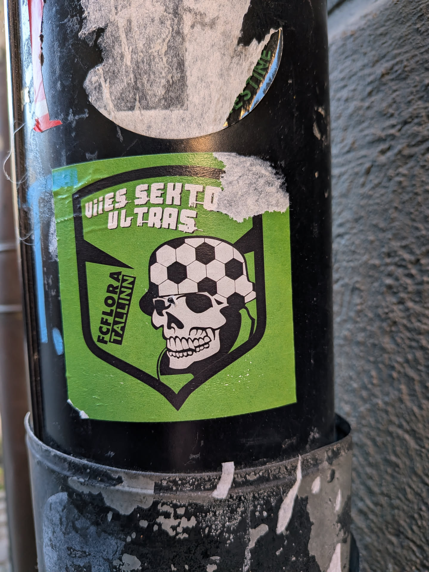 The sticker is a shield-shaped emblem, predominantly bright lime green with bold black outlines and details. It features a stylized white skull with black eye sockets and teeth, wearing a helmet patterned like a classic black and white soccer ball. Above the skull, within the shield, is the text "VIIES SEKTO ULTRAS" in white, with some letters showing signs of wear. Along the left side of the shield, oriented vertically, is "FC FLORA TALLINN" in white text. The sticker is applied to a black pole and exhibits signs of age and wear, including peeling edges and faded areas, particularly at the top and bottom.
