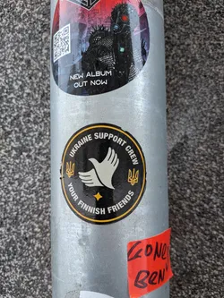 Street sticker A circular sticker with a black background and a bright yellow outer border. The central motif is a stylized white dove with outstretched wings, symbolizing peace. Below the dove, a small yellow four-pointed star is present. On either side of the dove, positioned towards the top of the circle, are two yellow Ukrainian trident symbols (Tryzub). Yellow capital letters arc along the top edge, reading "UKRAINE SUPPORT CREW", and along the bottom edge, stating "YOUR FINNISH FRIENDS". The sticker appears slightly worn, indicating real-world application.