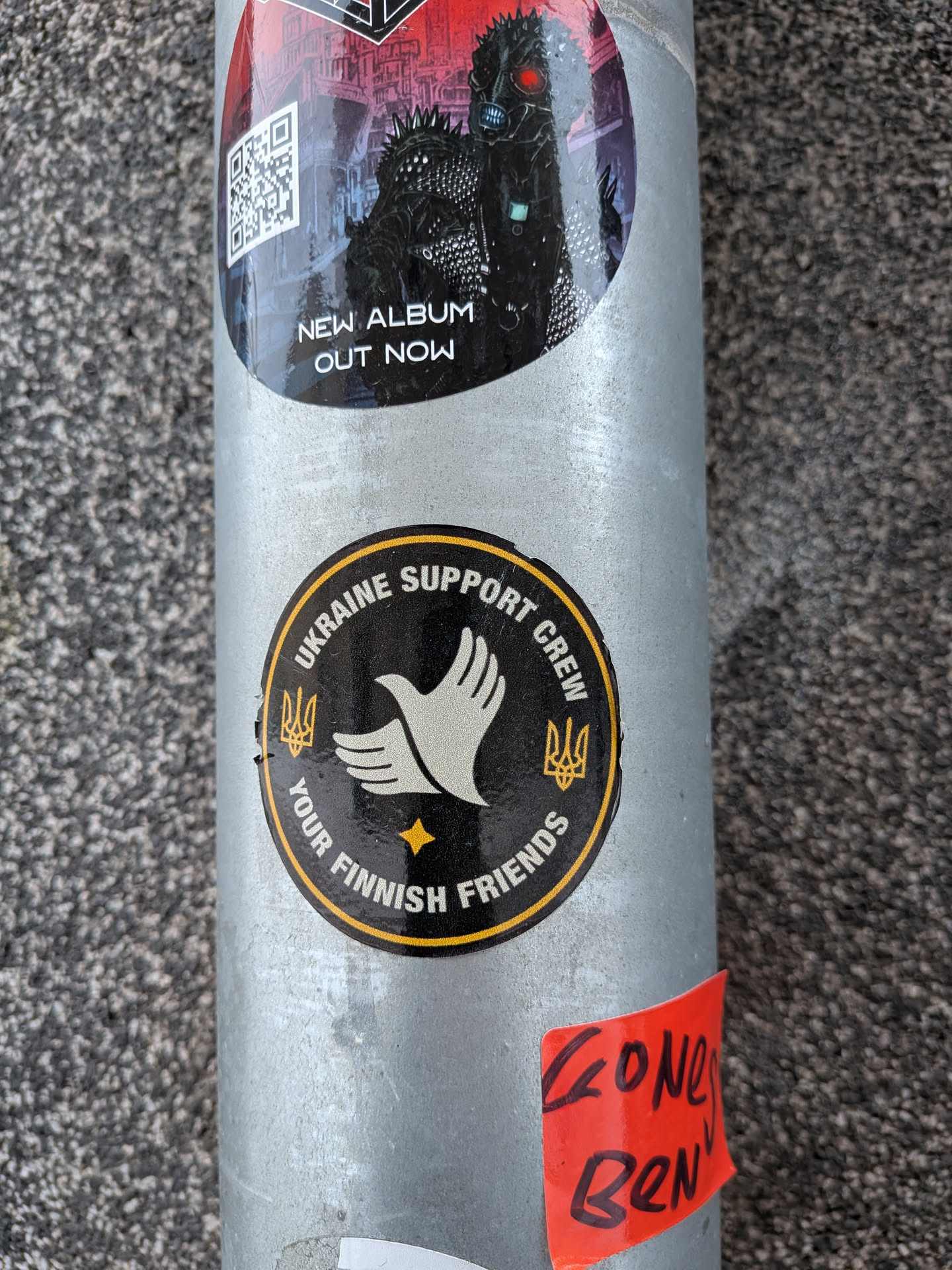 Street sticker A circular sticker with a black background and a bright yellow outer border. The central motif is a stylized white dove with outstretched wings, symbolizing peace. Below the dove, a small yellow four-pointed star is present. On either side of the dove, positioned towards the top of the circle, are two yellow Ukrainian trident symbols (Tryzub). Yellow capital letters arc along the top edge, reading "UKRAINE SUPPORT CREW", and along the bottom edge, stating "YOUR FINNISH FRIENDS". The sticker appears slightly worn, indicating real-world application.