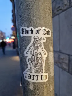 Street sticker A sticker featuring a black and white illustration of a skeletal figure with a snake, standing atop what seems to be a monstrous hand or creature.  The text "Mark of Cain" is at the top and "TATTOO" at the bottom, both in a gothic style font.