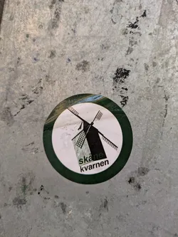 Street sticker This round sticker has a white background with a green border. A black windmill graphic is in the center, and below it, the words "Skåne kvarnen" are printed in black.