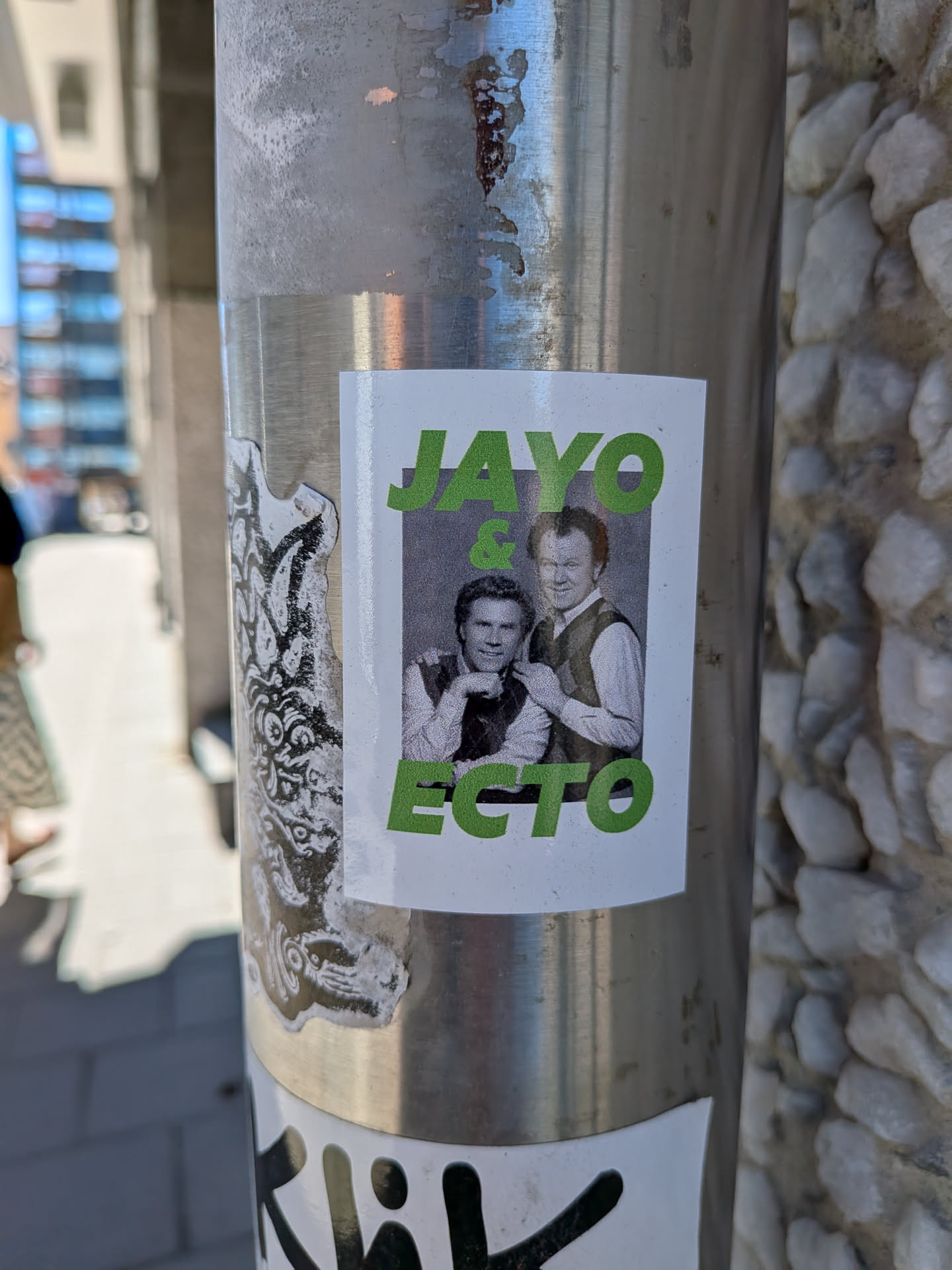 The sticker is rectangular with a clean white background. Centered on the sticker is a black and white photograph depicting two smiling men, who strongly resemble actors Will Ferrell and John C. Reilly. The man on the left (Will Ferrell) is leaning slightly forward with his hand near his chin, wearing a light-colored shirt and a dark vest. The man on the right (John C. Reilly) is positioned slightly behind and to his right, also smiling, wearing a similar light shirt and dark vest. Above the photo, in bold, slightly textured green capital letters, is the word "JAYO". Below the photo, in the same bold, textured green capital letters, is the word "ECTO". An ampersand "&" in the identical green font is placed between "JAYO" and the top edge of the photograph. The sticker is affixed to a metallic pole and shows minor signs of wear around its edges.