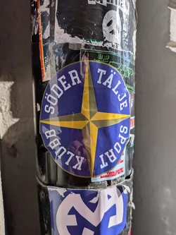 Street sticker A circular sticker featuring a dark blue background. At its center is a prominent four-pointed compass rose design in yellow, with a subtle darker yellow or gold outline. The points of the compass rose extend towards the edge of the circle. Arched around the top half of the circle, in bold white capital letters, is the text "SÖDER TÄLJE". Arched around the bottom half, also in bold white capital letters, is "SPORT KLUBB". The sticker has a glossy finish, reflecting some light, and appears slightly wrinkled, indicating it's a physical sticker adhered to a pole covered with other worn stickers.