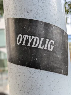 Street sticker A black and white sticker with the word "OTYDLIG" written in white capital letters on a black background. The sticker is affixed to a light-grey pole. The sticker is slightly dirty or worn.