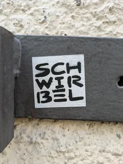 Street sticker A square, white sticker with black text is adhered to a gray metal fixture. The sticker reads 'SCHWIRBEL' in a stacked format. The text is bold and slightly distressed in appearance. The sticker is centrally located in the frame, with the metal fixture providing a contrasting backdrop. The fixture is attached to a textured, light-colored wall.